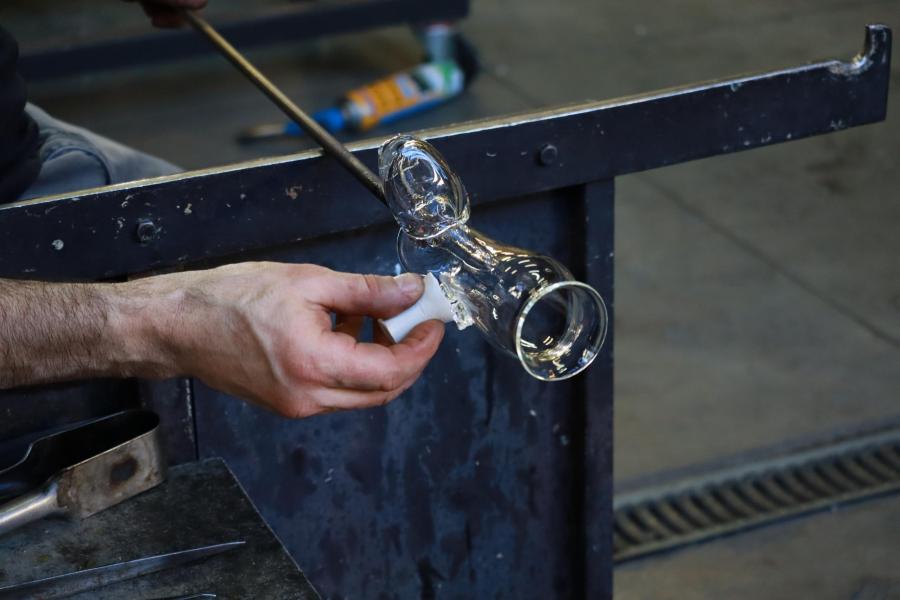 Een glaskunstenaar werkt in het atelier aan een transparant glazen drinkglas met vervormde, speelse vorm. Het stuk wordt aan een blaaspijp vastgehouden en met de hand verder gemodelleerd, als voorbeeld van trickware: glaswerk dat verrast door zijn ongewone vorm en functie.